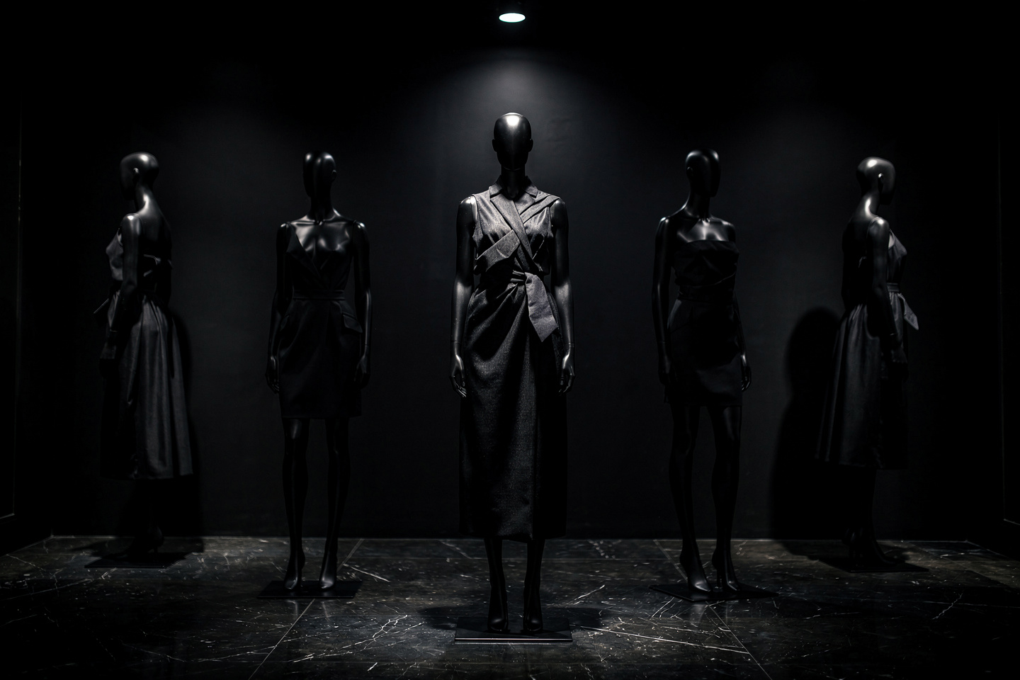 Editorial photo of an ultra-dark high-fashion boutique interior, moody black-on-black couture garments displayed on sculptural mannequins, dramatic low-key lighting with a single focused spotlight, deep shadows, matte black walls and polished dark marble floor reflecting subtle highlights, cinematic atmosphere, shot on 50mm lens, shallow depth of field, luxury minimalist composition, high contrast, sophisticated and mysterious mood.